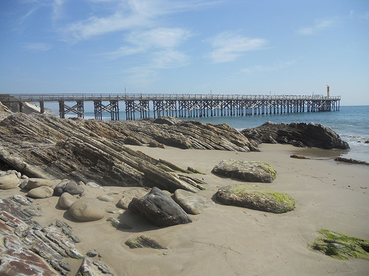 a wooden pier extends along the horizon, round moss covered and angular sandstone rocks extend from the sand meeting the ocean and pier 